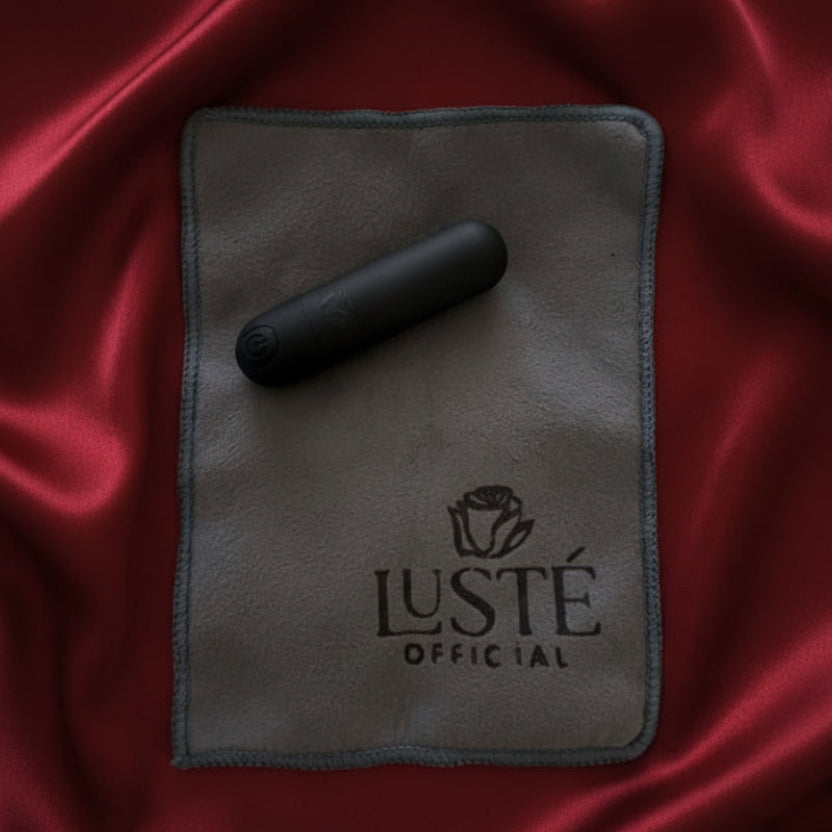 Black cylindrical vibrator on a gray cloth with 'Lusté Official' branding on a beige fabric background